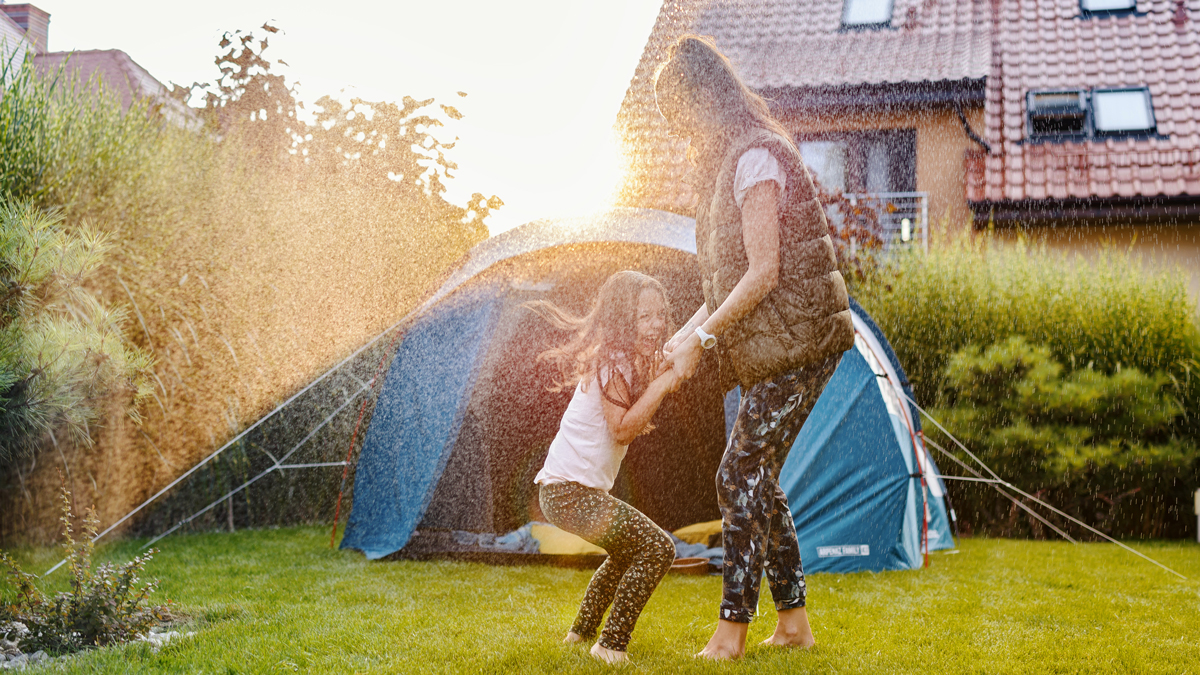 Mama und Tochter haben Spaß im Garten im Rasensprenger mit Zelt im Hintergrund