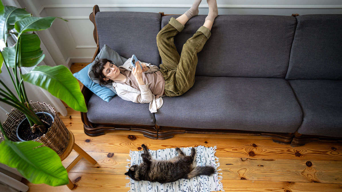 Couchsurfing vs Untervermietung, Frau chillt auf dem Sofa mit Smartphone in der Hand, davor liegt eine Katze auf dem Boden und schläft