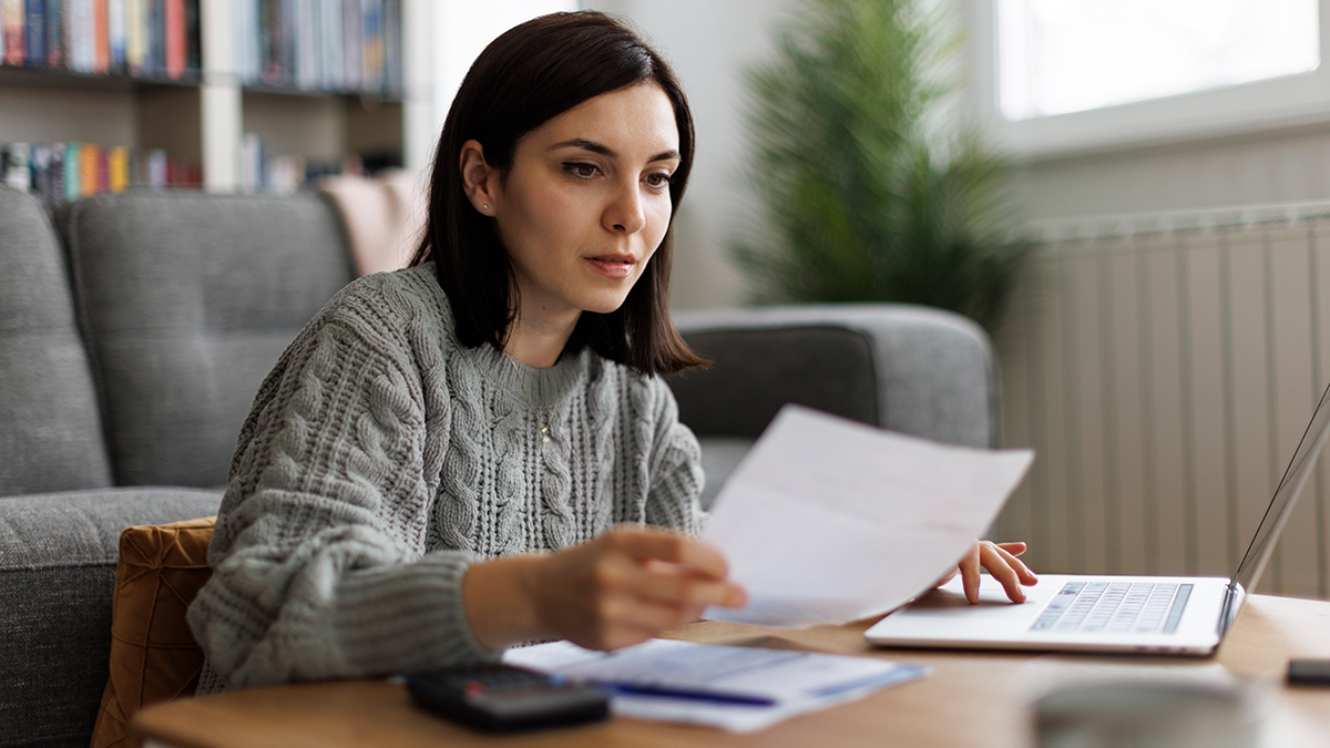 Eine Frau am Laptop schaut auf ihren Einkommensnachweis in der Hand