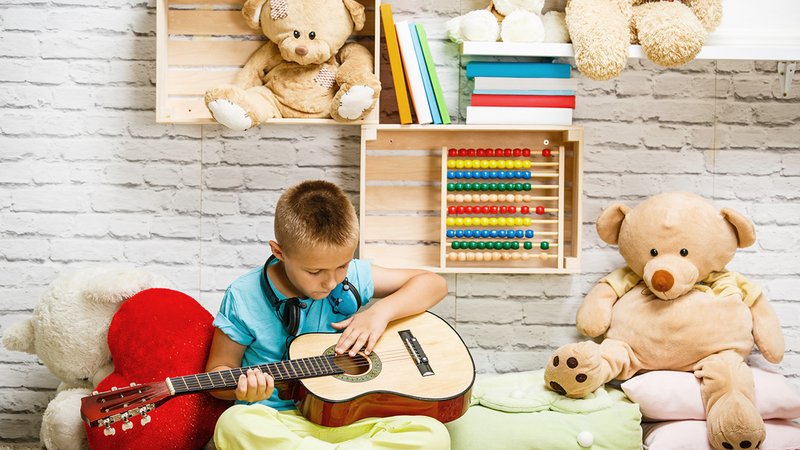 Upcycling Kinderzimmer, ein Junge sitzt in seinem Kinderzimmer und spielt Gitarre vor einem Spielzeugregal aus Holzkisten