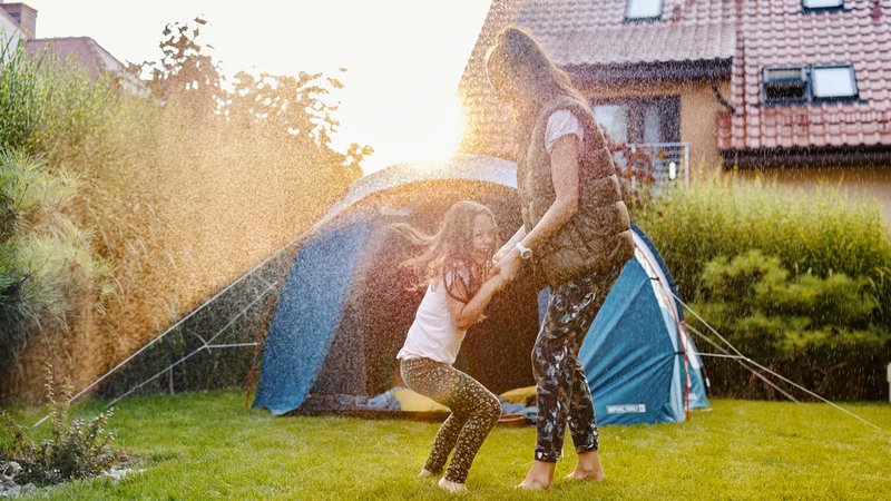 Mama und Tochter haben Spaß im Garten im Rasensprenger mit Zelt im Hintergrund