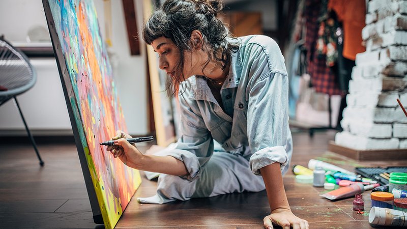 Alleine wohnen Vorteile, Frau sitzt am Boden in ihrer Wohnung und malt ein Bild