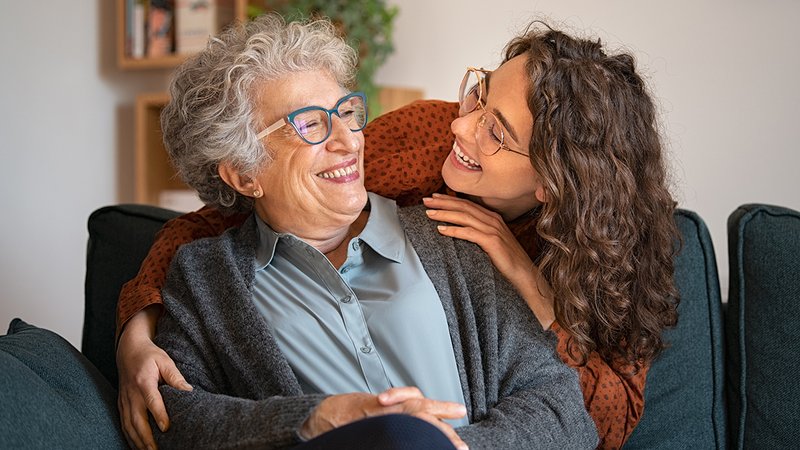 Haus verkaufen mit Wohnrecht, ältere Frau sitzt auf Sofa und wird von einer jüngeren Frau von hinten umarmt, Foto: Rido / stock.adobe.com