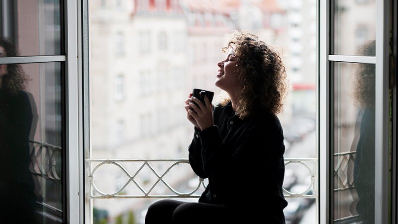 PM AT Sicherheit, Frau trinkt zufrieden einen Kaffee vor offenem Fenster