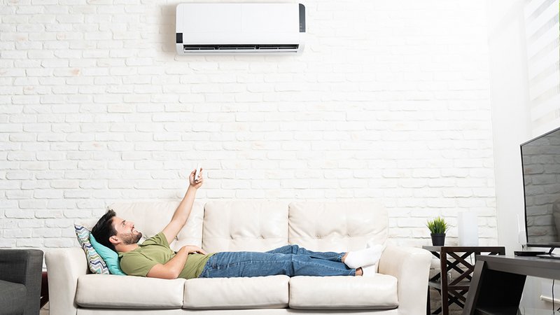 Klimaanlage dynamischer Stromtarif, Mann liegt entspannt am Sofa und schaltet mit Fernbedienung die Klimaanlage ein