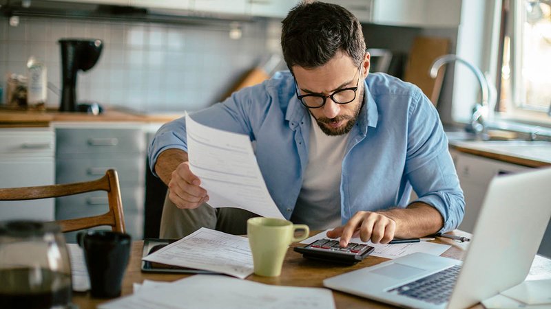 Mann sitzt mit Dokumenten und Laptop an einem Küchentisch